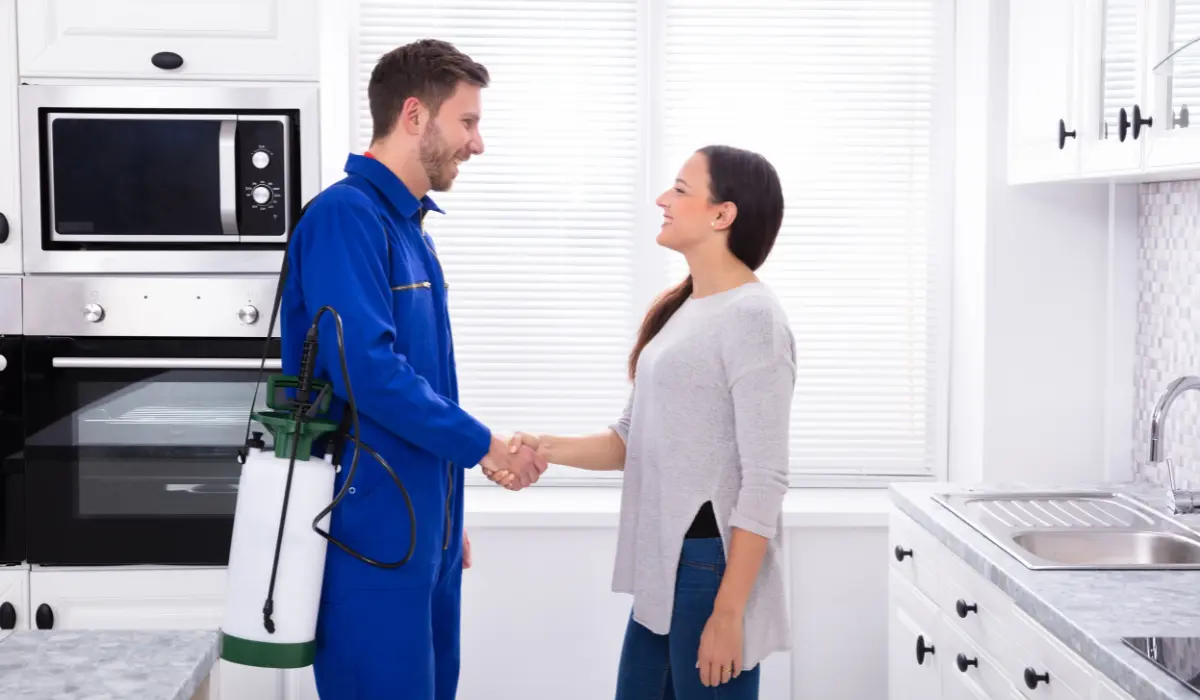 Pest control technician in blue uniform shaking hands with a smiling homeowner in a bright white kitchen