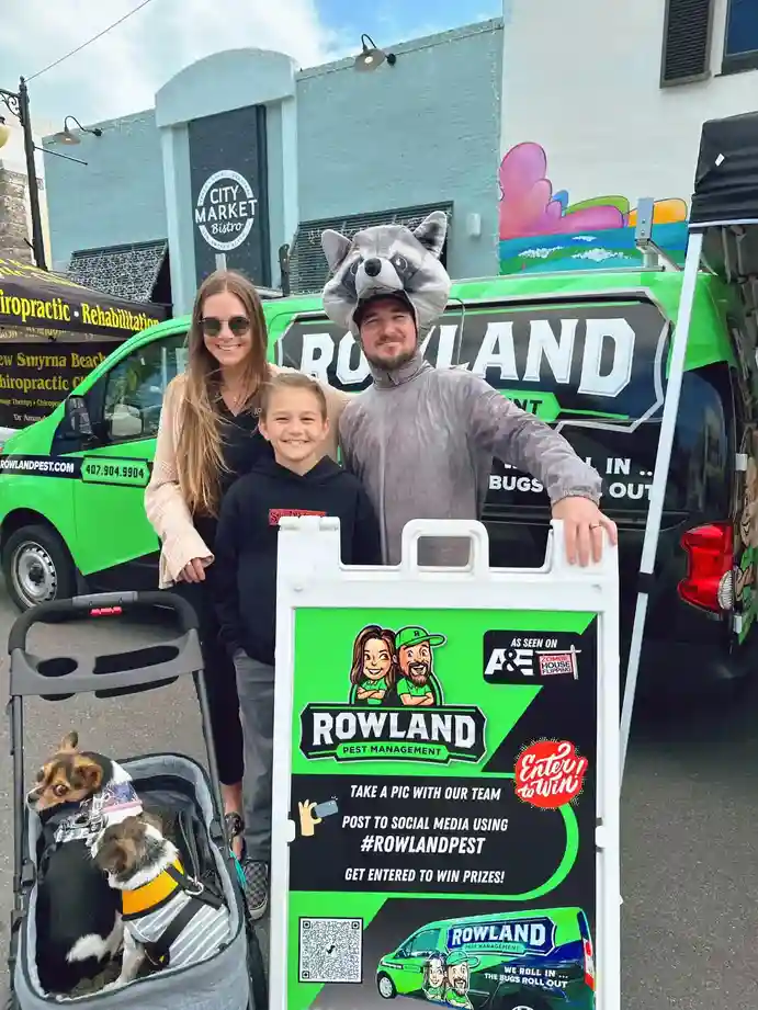Woman, child and person in raccoon costume pose by a green Rowland Pest Management van and sign, two small dogs in a stroller